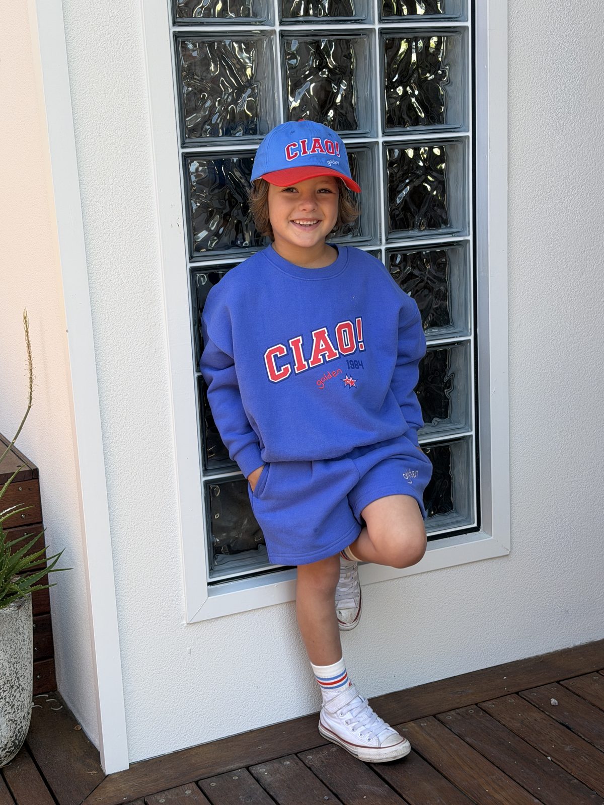 Boy in blue graphic sweatshirt and cap smiling by a glass block window
