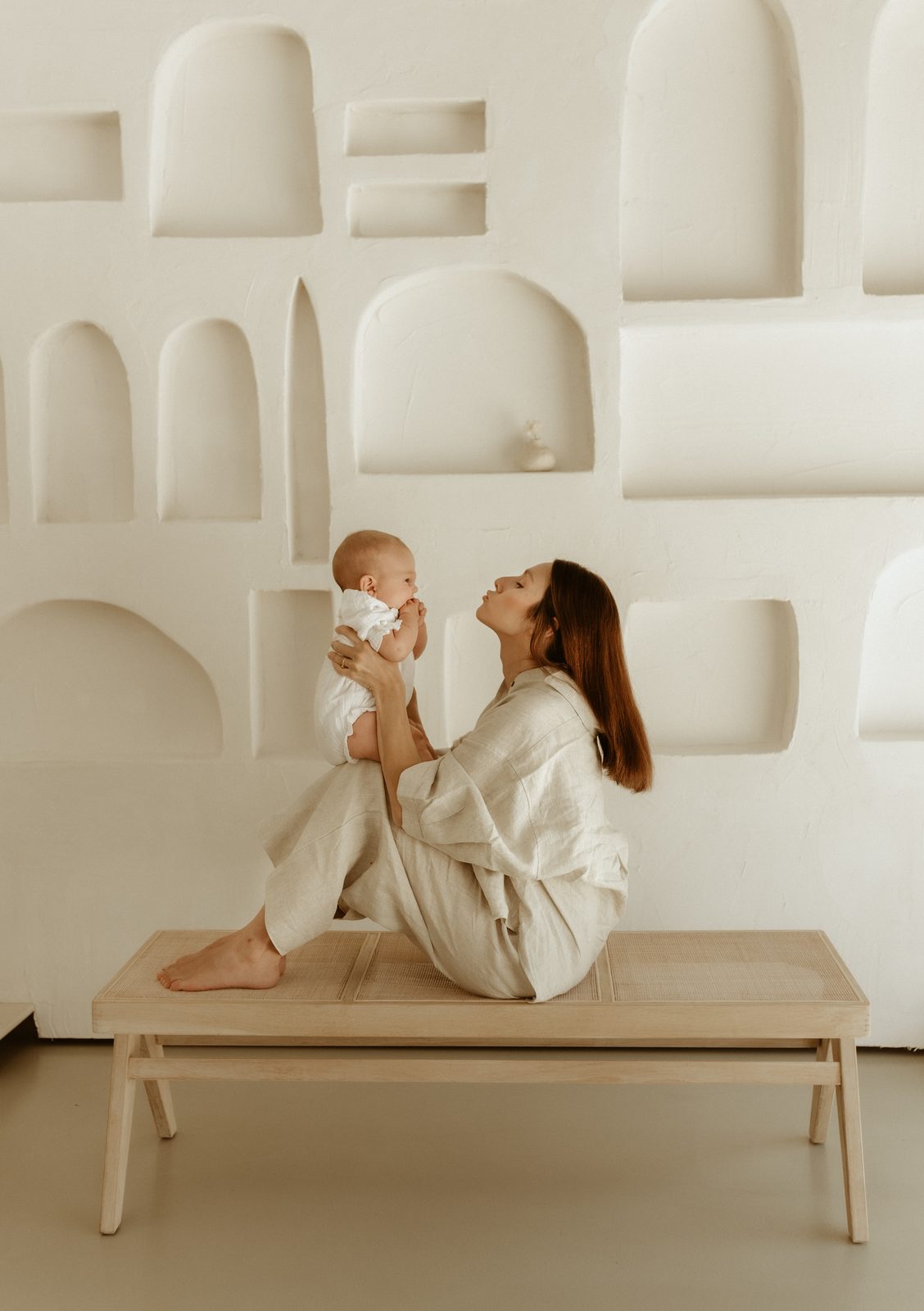 Mother in linen holding her baby up playfully in a sculptural white studio