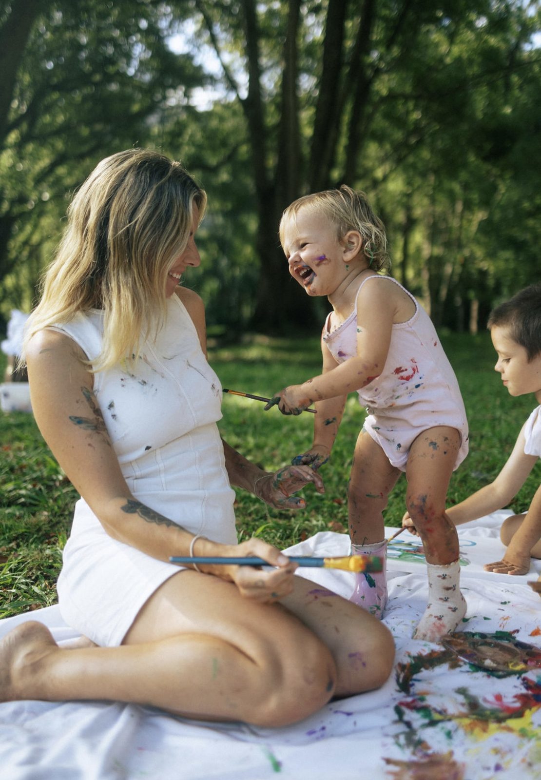 Mother laughing with paint-covered toddler and child during an outdoor painting session